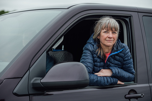 A shot of a mature adult female with a fringe leaning out of a van window. She has a neutral expression, with her arms crossed, and is wearing a padded jacket. The van is parked at the coast in Alnmouth, North East England.