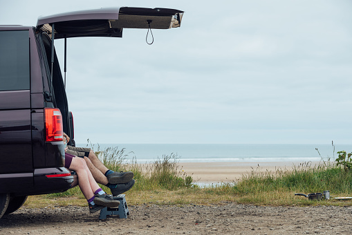 A side-view shot of a sea view with camper van parked up in partial view to one side. There are two people sitting in the trunk of the van, both dangling their feet out of the back, in Alnmouth, North East England. There is green foliage lining the edge of the gravel car park, behind which the sand and sea are visible.

Videos available of this scenario.