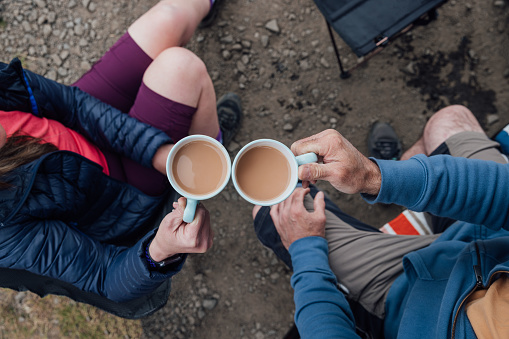 An overhead shot of two unrecognisable people holding mugs of tea whilst sitting outside on camping chairs at the coast in Alnmouth, North East England. They are clinking their mugs together in a celebratory toast. They are both wearing sportswear and a warm jumper or jacket suitable for a British summer.