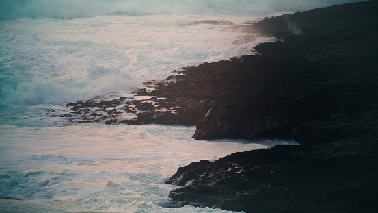 Stormy sea hitting rocky coastline in evening. Powerful waves rolling breaking volcano stones in early morning. White foaming surf washing beach in slow motion. Dramatic seascape nature landscape.