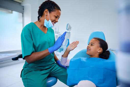 Happy African American little girl and her dentist giving high five after teeth examination at dentist's office.