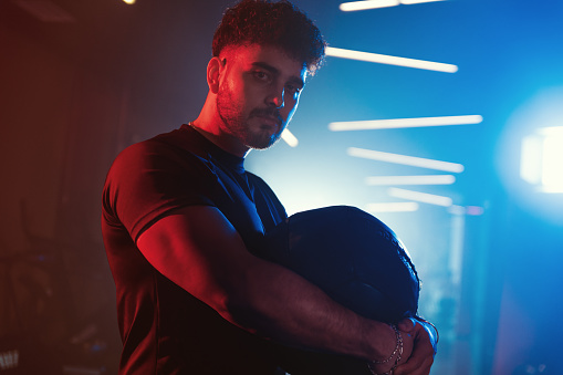 Portrait of a male athlete holding a medicine ball, captured under the neon blue and red lights of a misty gym