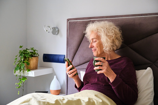 Mature Hispanic woman using the smartphone and drinking coffee on her bed.