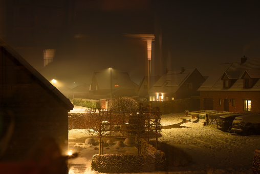 Leuven, Flemish-Brabant, Belgium-January 18, 2024: high angle view reflections scene from a window first story. Snow falling at evening on a suburb of Leuven. A thick layer of snow lies on the roofs. Snow lit up under lampshade street lights