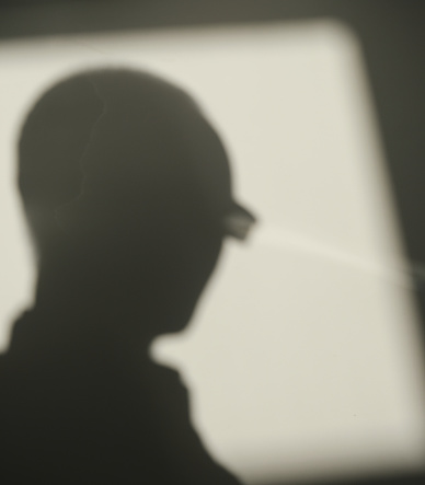 Sun lit shadow of a bald adult male head and shoulders (wearing glasses) in profile. Dark, mysterious, moody detective noir retro movie style. Isolation, depression or loneliness. Close up copy space