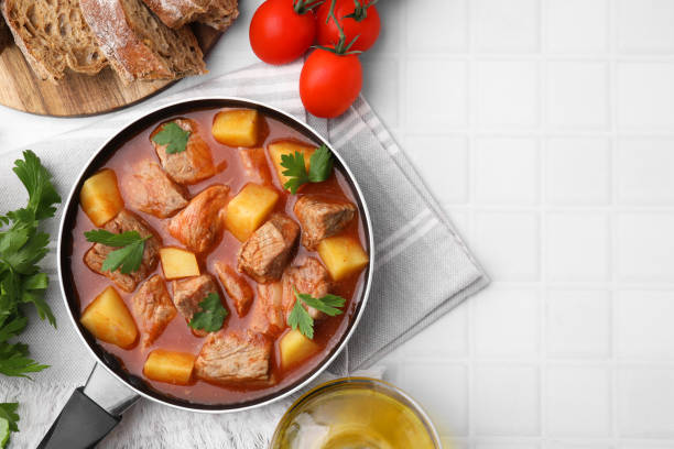 istock Delicious goulash in saucepan and ingredients on white tiled table, flat lay. Space for text 1942774947