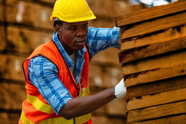istock African worker carpenter wearing safety uniform and hard hat working and checking the quality of wooden products at workshop manufacturing. man and woman workers wood in dark warehouse industry. 1942787699