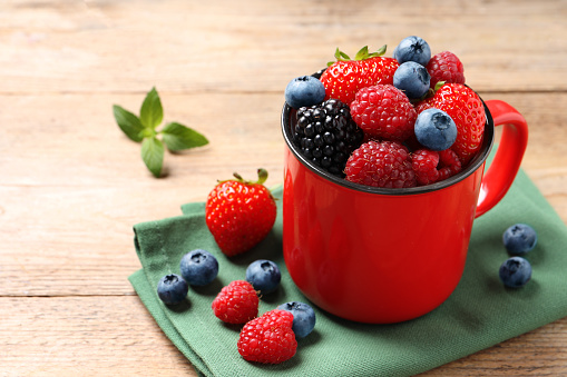Many different fresh ripe berries in mug on wooden table, space for text