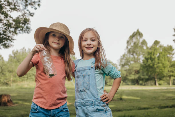 Adorable cheerful little twin girls with long dark hair in similar white dresses and straw hats smiling brightly while playing together on grassy meadow during holidays in countryside