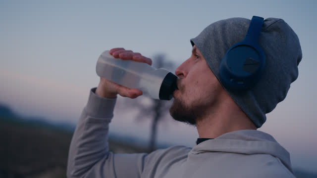 SLO MO Closeup Side View of Young Male Athlete Wearing Headphones while Drinking Water from Bottle in Countryside During Blue Hour