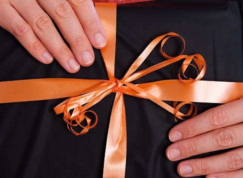 A close-up shot of a black gift with an orange bow in woman hands.
