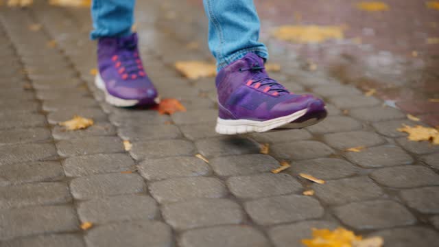 closeup female legs in sport shoes, sneakers. woman walking at rainy autumn day in park. Rain drops Falling On puddle with yellow maple leaves reflection on city street sidewalk