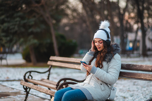 Young woman sitting on a park bench while listening to music.