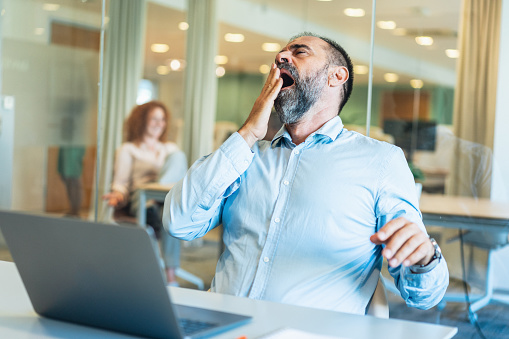 Overworked mature businessman yawning tired of work on laptop sitting at desk in the modern office. Exhausted mature businessman yawning. Senior businessman spent many hours working.