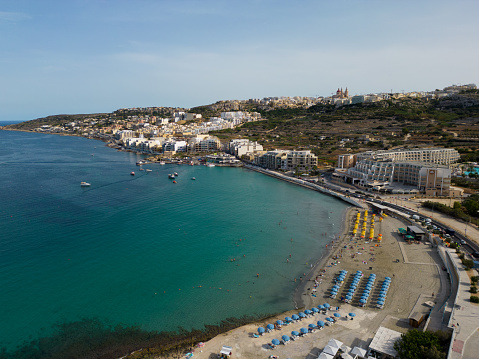 Ghadira Bay with sandy beach and sun umbrellas. Mellieha coastal town next to the beach in the summer Malta aerial.
