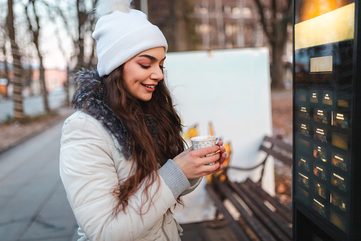 Beautiful young woman buying coffee outdoors on a coffee vending machine.