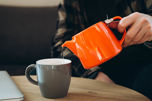 A man pours tea from a bright orange teapot into a cup. A man drinks hot tea in the cold season. Tea break at work.