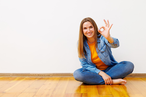 Smiling woman in jeans and denim jacket sitting on the floor giving an OK hand sign, expressing satisfaction.