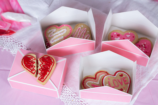 Boxes of heart shaped cookies are sitting together on a table. Three of the boxes are open, showing the sugar cookies decorated with colored icing and sprinkles. One box is closed, with a broken cookie on top; the broken cookie is decorated like a broken heart.