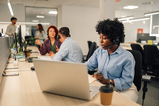 Portrait of an african american woman with headset working on a laptop in an open space office. Call center operator with wireless headset talking with customer and resolving issues. Copy space.