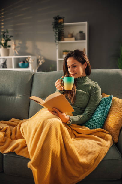 istock Woman drinking tea while reading a book on a sofa at home covered with blanket 1943155164