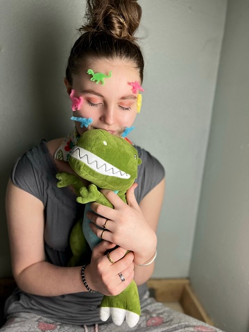 Autistic beautiful teen girl who loves dinosaurs. She is displaying her love and hobby by placing foam dinosaur cut outs on her face and holding and kissing a stuffed dinosaur. Her eyes are closed and she is embracing her stuffed animal.