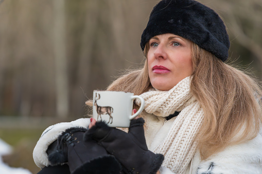 Close-up of a woman with a fur hat holding a coffee mug, gazing into the distance, on a cold winter day