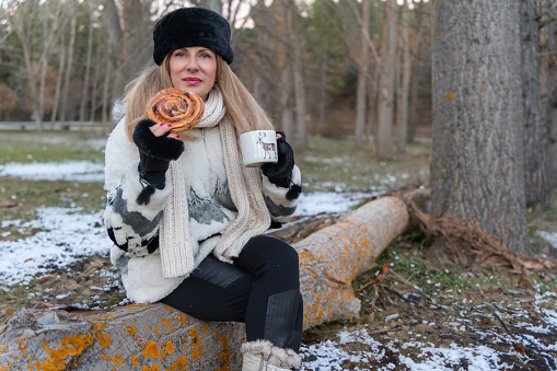 Smiling woman in a fur hat and winter coat enjoys a pastry and hot drink while seated on a snow-dusted log