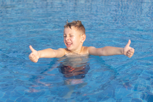 Little boy wearing a blue uv swimsuit and blue swimming goggles is smiling as he is learning to swim holding a noodle float in an outdoor swimming pool in summer. This is a close-up picture of the swimming child only.