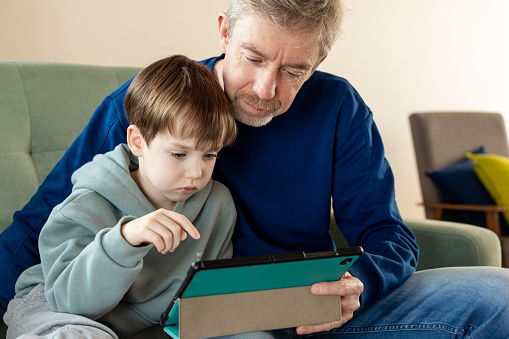 A 6 year old boy and his dad are relaxing on the sofa and using digital devices