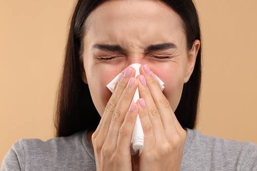 Suffering from allergy. Young woman blowing her nose in tissue on beige background, closeup