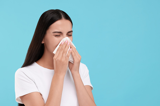 Suffering from allergy. Young woman blowing her nose in tissue on light blue background. Space for text