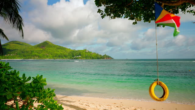 handheld shot of a empty tropical beach in the Seychelles Islands, Indian Ocean. View of national flag waving on top of a yellow tire tired on a rope. View of the exotic tropical white sanded beach.