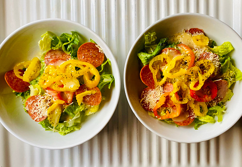 Pepperoni and Banana Peppers Salad with lettuce and Parmesan cheese in white bowls on a kitchen counter top near a window with natural light.