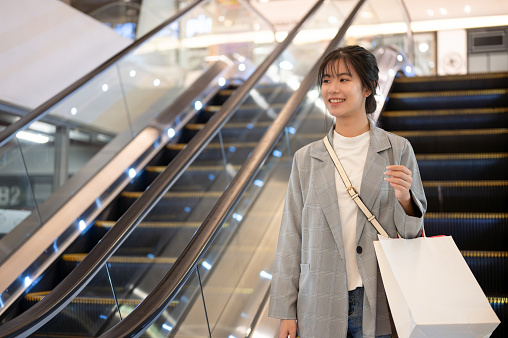 An attractive young Asian woman in trendy clothes with shopping bags is going down an escalator in a shopping mall. city life, consumerism, lifestyle concepts