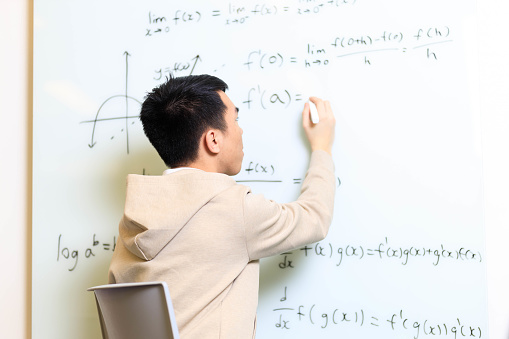 An Asian male young adult teenage student is writing on a glass whiteboard filled with math equations in a university or high school interactive learning classroom. He is wearing a beige hooded jacket and a white polo shirt.