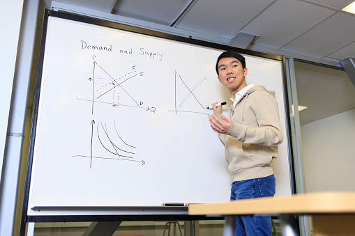 An Asian male young adult is holding up a marker in front of a whiteboard tutoring economics in a university or high school interactive learning classroom. He is speaking to the audience. He is wearing a beige hooded jacket and a white polo shirt.