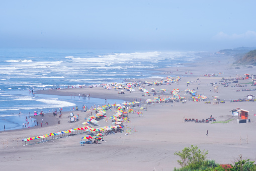 Top view at famous Parangtritis Beach, Yogyakarta, Indonesia. Colorful tents, vehicles, hut and crowded of people swimming, relaxing, walking along white sandy shoreline with blue water.