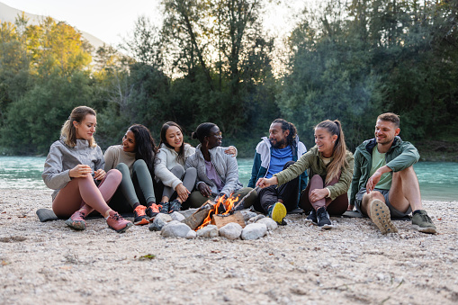 Diverse group of friends sitting by a campfire near a river, enjoying a casual outdoor gathering.