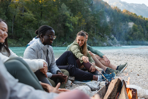 Group of friends from diverse ethnic backgrounds laughing and relaxing by a campfire near a river with mountains in the background.