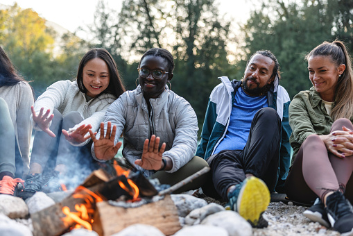 A group of diverse friends warmly dressed, gather around a campfire, sharing a convivial moment in an outdoor setting.