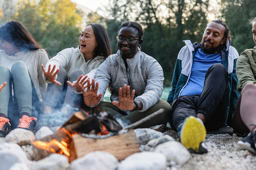 A group of diverse friends warms their hands over a cozy campfire, sharing stories and laughter in an outdoor setting.
