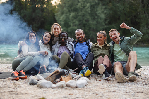 Group of joyful friends of various ethnicities sitting by the campfire, sharing laughter and stories in a natural outdoor setting.