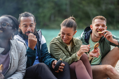 Group of diverse friends share laughs and snacks while sitting by a campfire outdoors, exemplifying friendship and leisure.