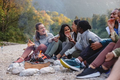 Friends of diverse ethnic backgrounds share laughter and food around a campfire in a natural outdoor setting.