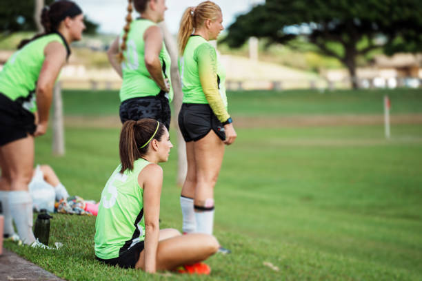 A young adult female athlete sits on the grass on the sideline with her teammates during a competitive soccer game on a beautiful day in Hawaii.