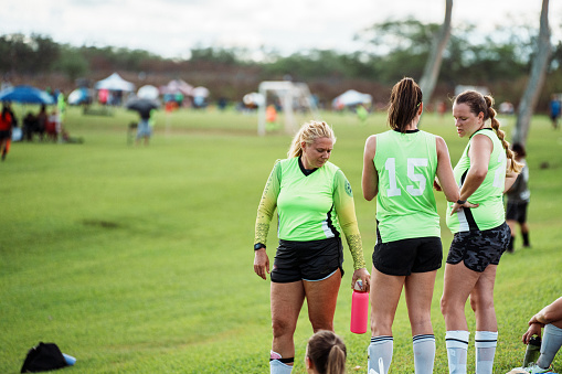 Young adult female athletes stand on the sideline with their teammates during a competitive soccer game in a club tournament.