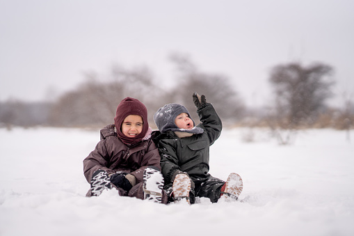 Two young siblings sit together in the snow on a cool winter day.  They are each dressed warmly in a snowsuit, hat, mittens and boots as they laugh with one another.