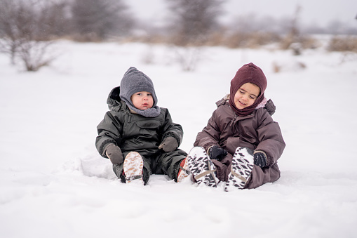 Two young siblings sit together in the snow on a cool winter day.  They are each dressed warmly in a snowsuit, hat, mittens and boots.