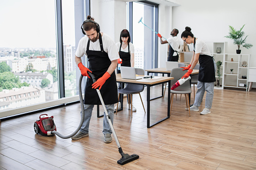 Team of young multicultural cleaners vacuums floor, wipes tables with gadgets, shelves, washes windows in spacious, bright, modern office. Professional cleaning company.